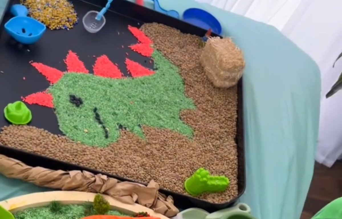 A dinosaur shaped from dyed green and red rice on a sensory tray, with scoops and a toy dinosaur alongside.