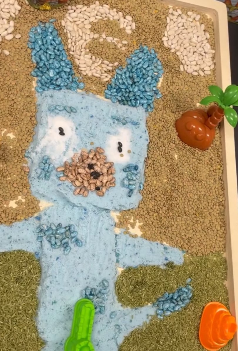 A themed sensory tray built from dyed rice, lentils and beans arranged as a character design.